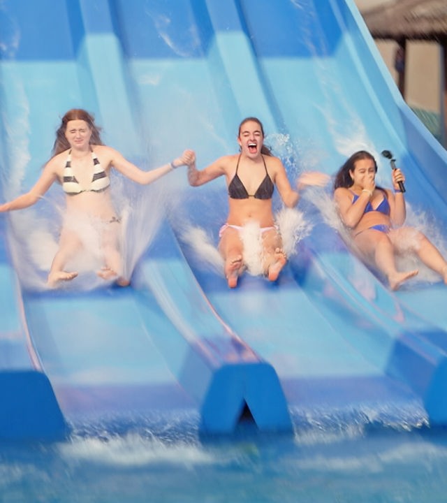 v.l.n.r. Marie, Lenja und Lina-Marie rutschen auf einer blauen Reihen-Wasserrutsche nebeneinander runter. Sie halten sich gegenseitig an den Händen, Lina-Marie hat eine GoPro in der freien Hand.