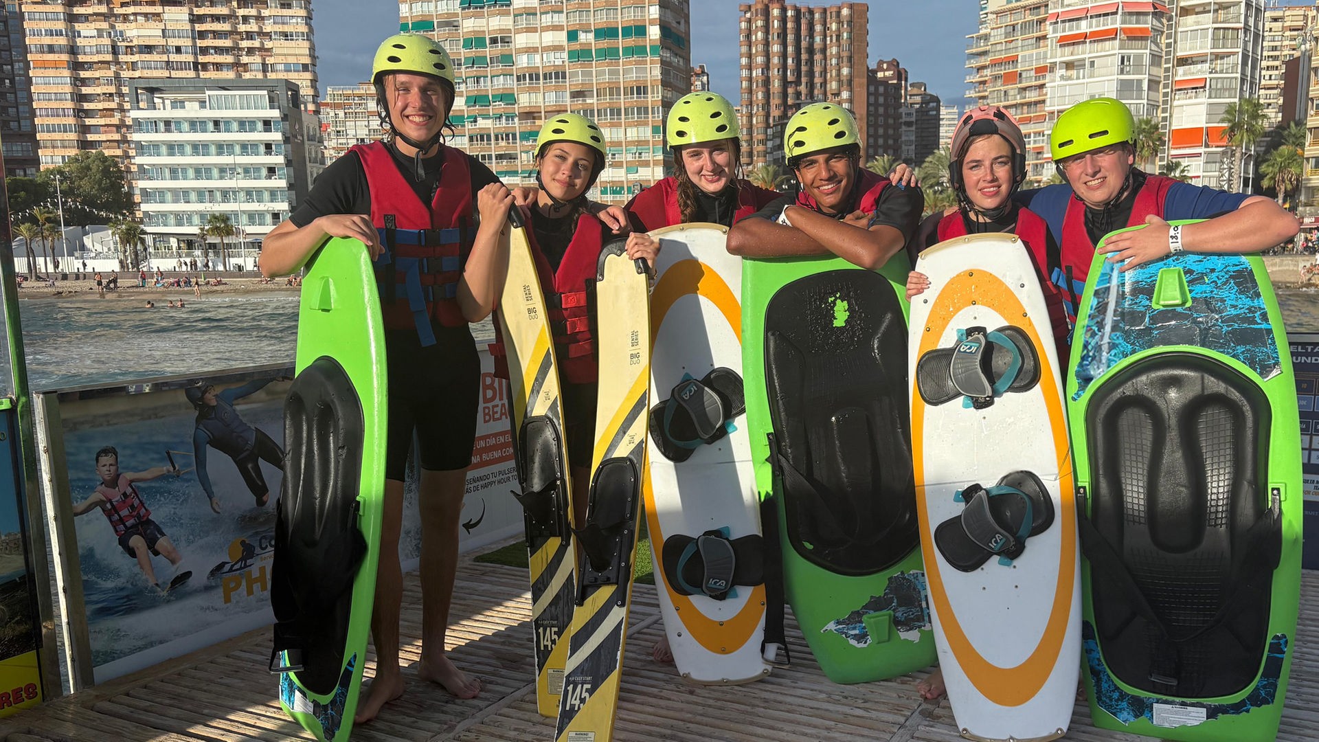 v.l.n.r. Jan, Lina-Marie, Marie, Adrian, Lenja und Rufus stehen auf einer Plattform und haben vor sich jeweils Wakeboards und Wasserski, auf die sie sich abstützen. Alle tragen Helme und lächeln in die Kamera. Im Hintergrund ist der Strand zu sehen.