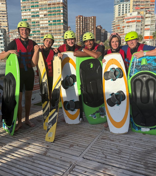 v.l.n.r. Jan, Lina-Marie, Marie, Adrian, Lenja und Rufus stehen auf einer Plattform und haben vor sich jeweils Wakeboards und Wasserski, auf die sie sich abstützen. Alle tragen Helme und lächeln in die Kamera. Im Hintergrund ist der Strand zu sehen.