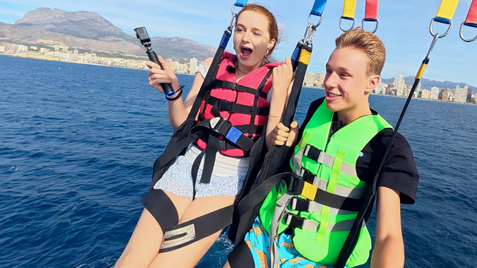 Marie links und Jan rechts hängen zusammen an einem Fallschirm beim Parasailing. Jan lächelt, während Marie eher erschrocken schaut. Sie sind kurz vor der Landung im Wasser.