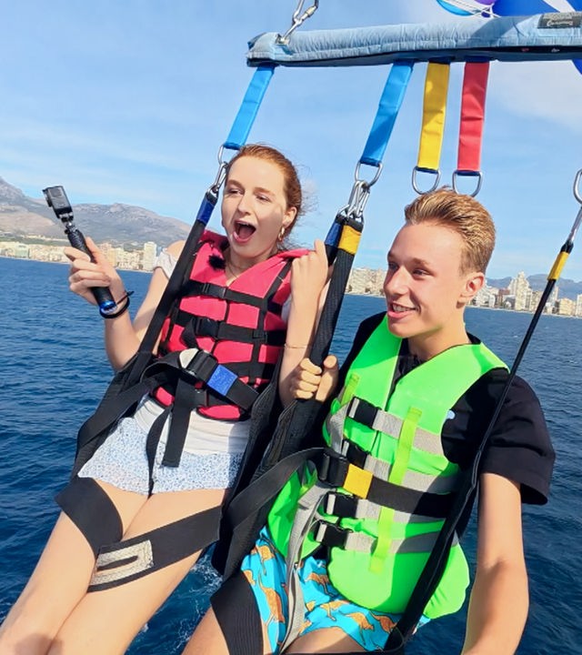 Marie links und Jan rechts hängen zusammen an einem Fallschirm beim Parasailing. Jan lächelt, während Marie eher erschrocken schaut. Sie sind kurz vor der Landung im Wasser.
