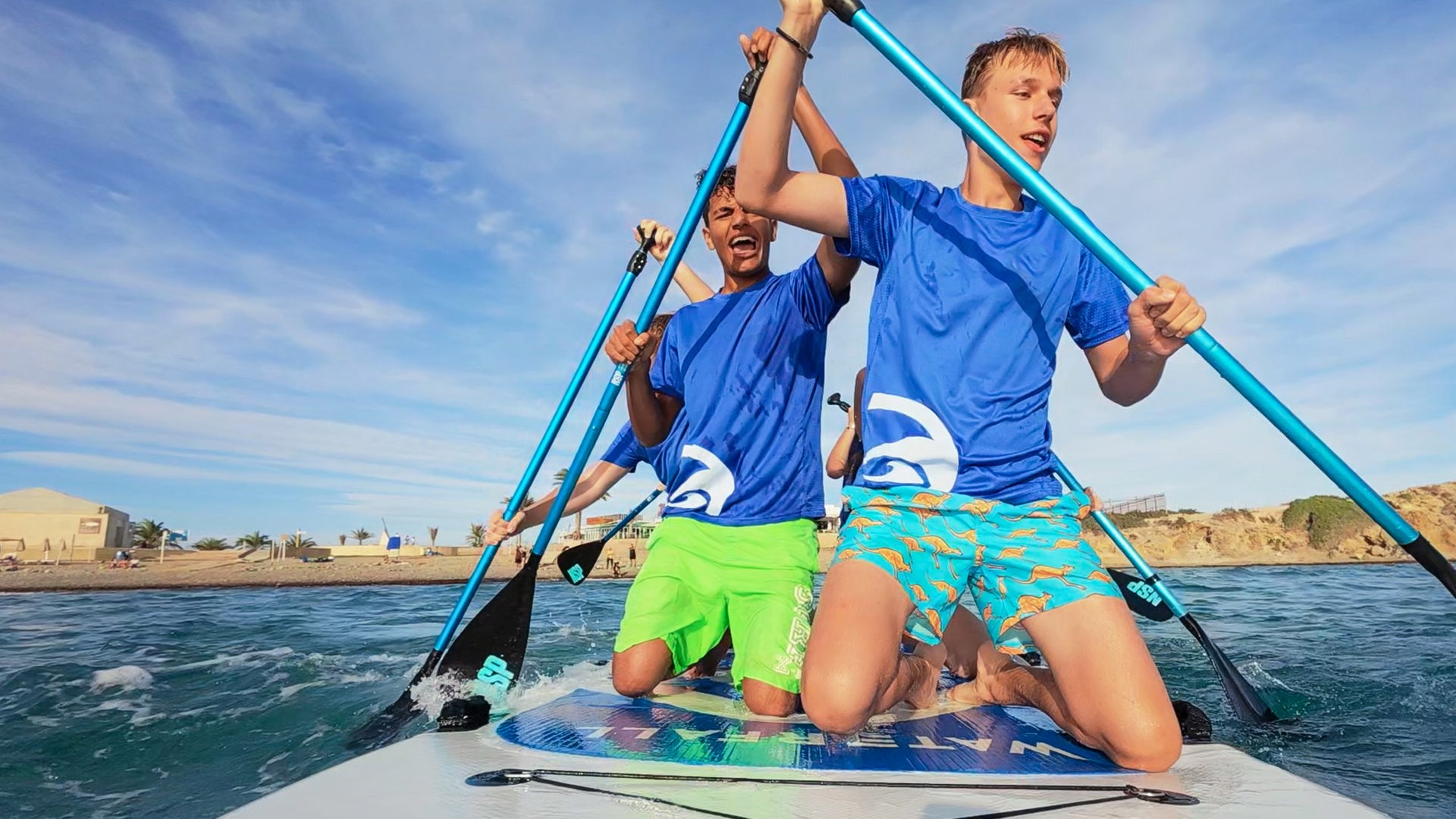 Jan kniet vor Adrian und weiteren Jugendlichen, deren Gesichter von Jan und Adrian bedeckt sind, auf einem Stand Up Paddling Board auf dem Wasser. Alle rudern fleißig.