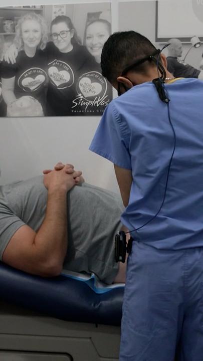 Ein Mann liegt auf einer Liege, man sieht seinen Oberkörper und sein Gesicht, sein Unterkörper wird verdeckt durch einen Mann in OP-Kleidung, der sich über den Mann auf der Liege beugt.