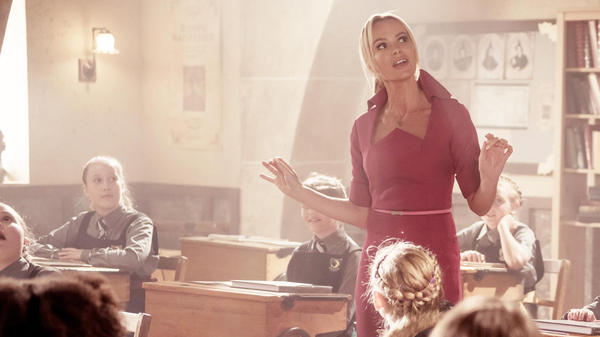 Frau Fünfzack steht mitten im Klassenzimmer. Die Schülerinnen sind begeistert und starren sie mit offenen Mündern und überraschten Gesichtern an.