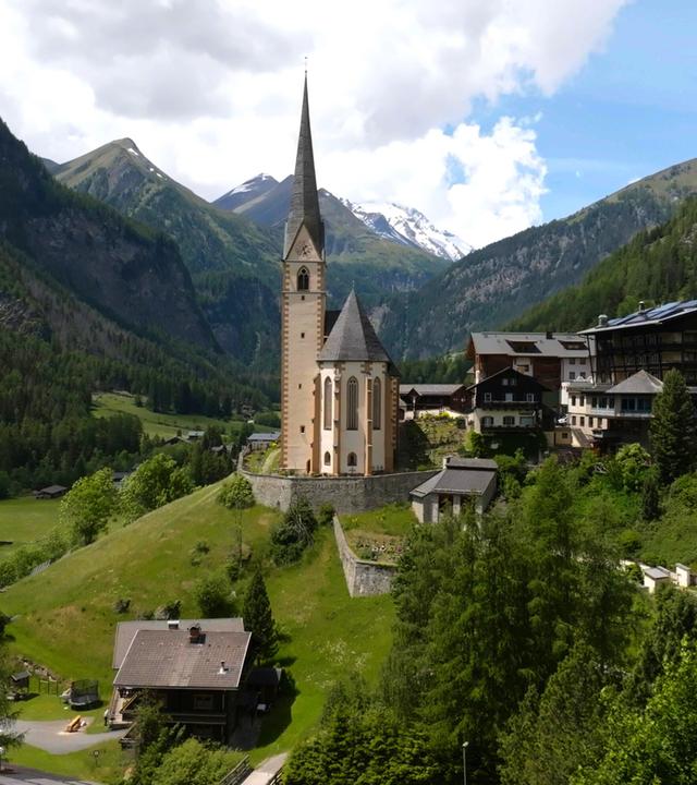 Das Bild zeigt eine malerische alpine Landschaft mit einer charakteristischen Pfarrkirche im Vordergrund. Die Kirche hat einen hohen, spitzen Turm und ist aus hellen Steinen erbaut, mit einigen dunklen Dachelementen. Im Hintergrund sind dramatische Berggipfel zu sehen, die teilweise schneebedeckt sind. Die Landschaft um die Kirche ist grün und bewaldet, mit sanften Hügeln und vereinzelten Häusern im Vordergrund. Diese Hütten sind traditionell aus Holz gebaut und haben Steildächer. Der Himmel ist teilweise bewölkt, was dem Bild eine dynamische Lichtstimmung verleiht. Diese Szene vermittelt ein Gefühl von Ruhe und Naturnähe in den Alpen.