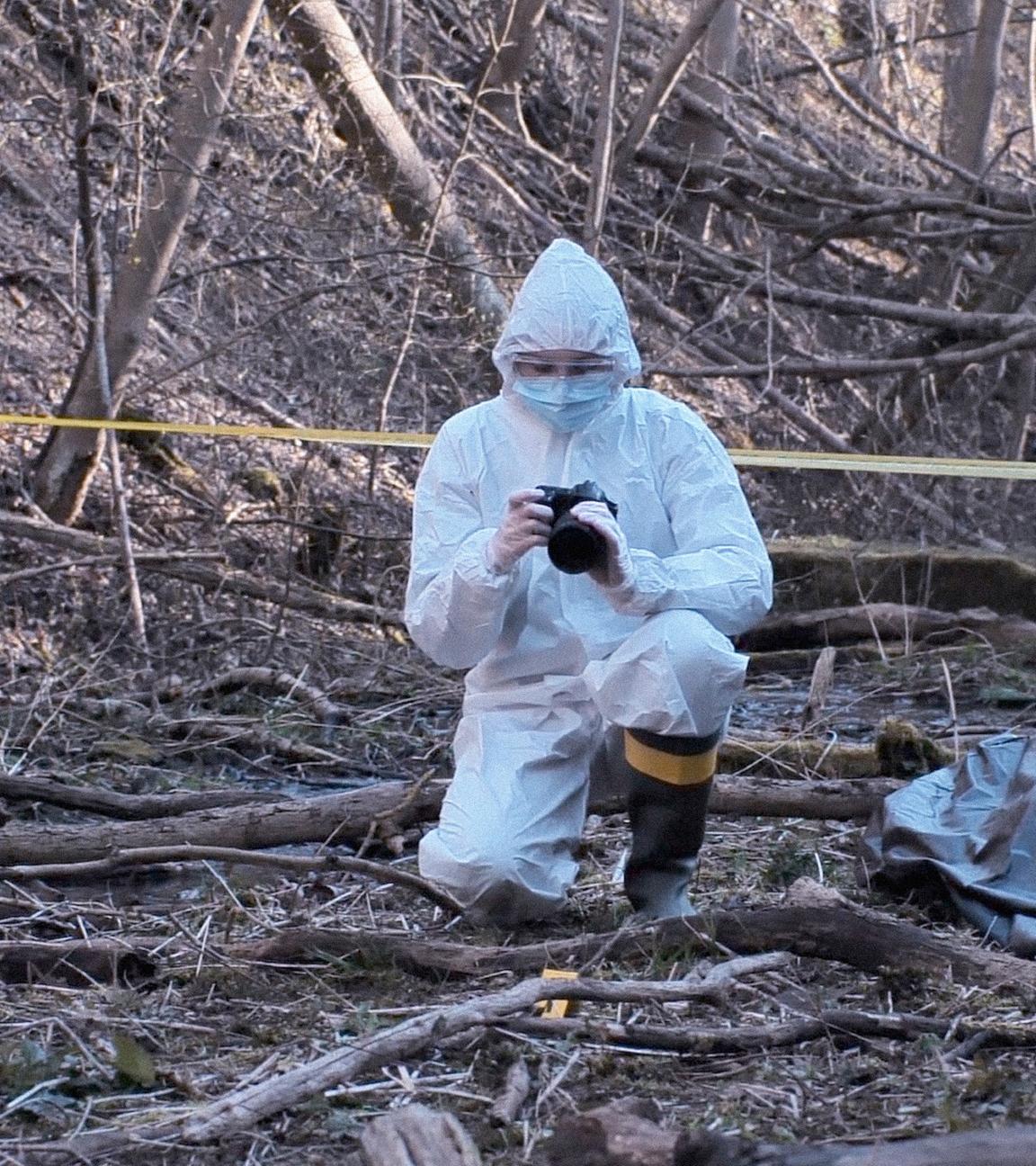 Zwei Männer in weißen Schutzanzügen sichern einen Tatort im Wald und machen Fotos.