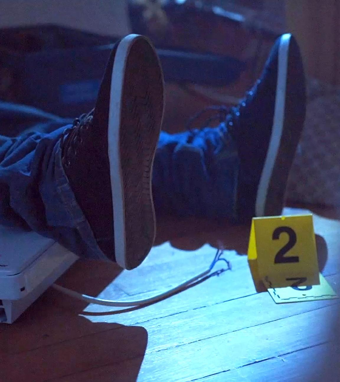Zwei Beine eines Mannes liegen auf dem Boden. An seiner linken Fußsohle steht ein kleines, gelbes Schild mit der Nummer 2.