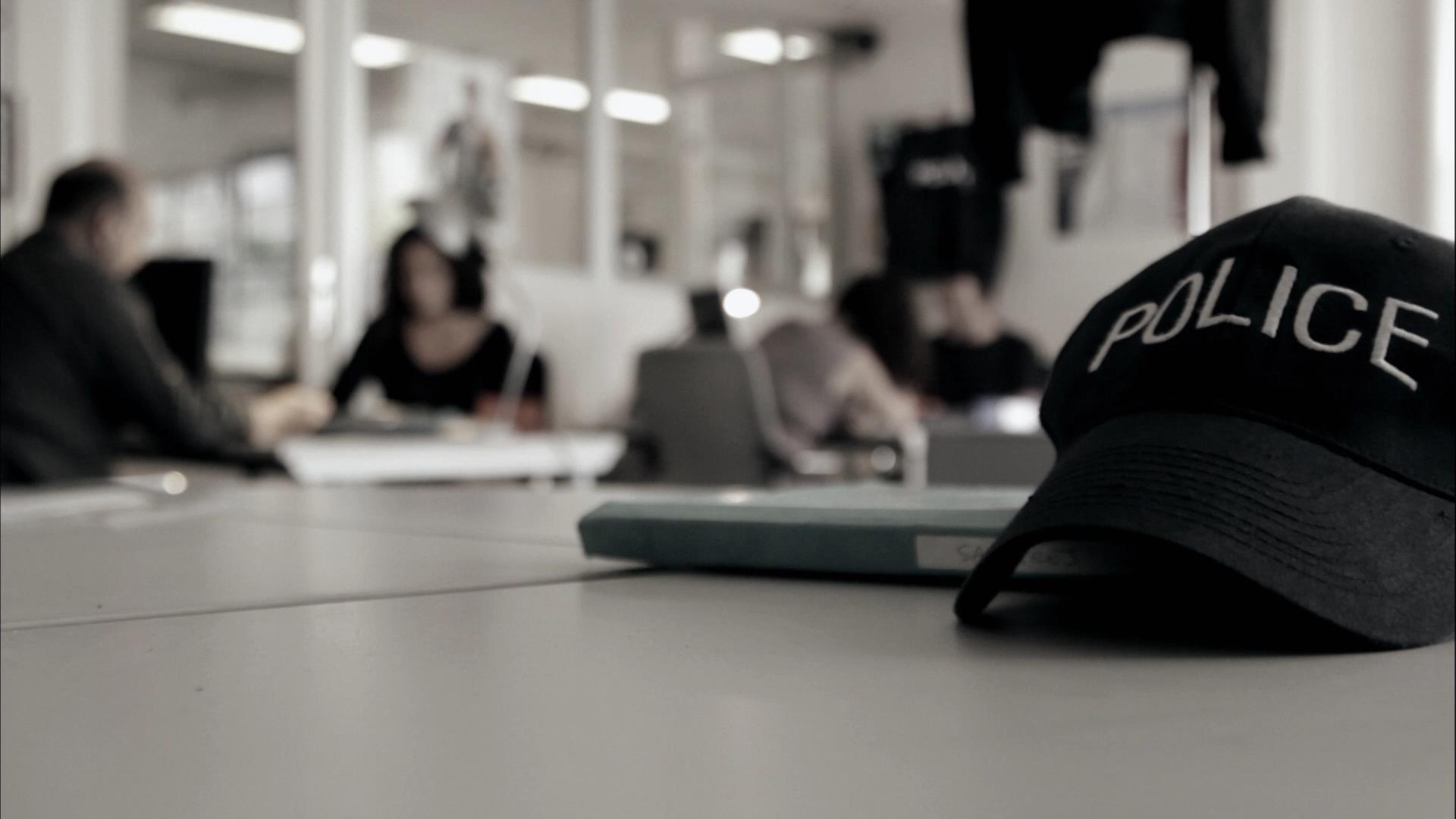 "Mörderjagd: Auf der Flucht": Eine Polizei-Kappe mit der Aufschrift "POLICE" liegt auf einem Tisch. Im Hintergrund ist verschwommen ein Büro einer Polizeistation mit mehreren Personen zu sehen.