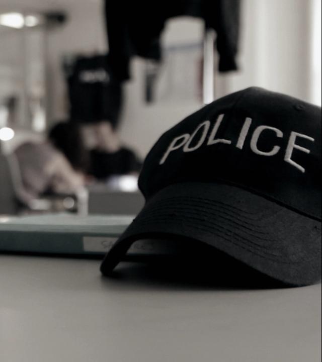 "Mörderjagd: Auf der Flucht": Eine Polizei-Kappe mit der Aufschrift "POLICE" liegt auf einem Tisch. Im Hintergrund ist verschwommen ein Büro einer Polizeistation mit mehreren Personen zu sehen.