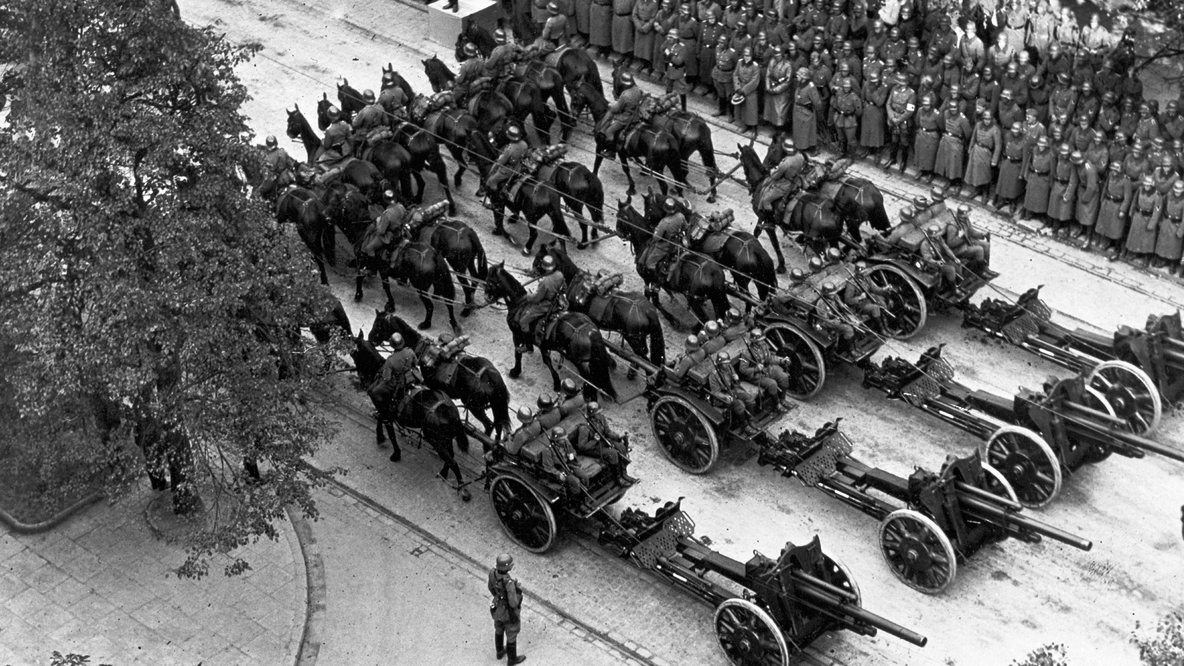 Die Deutsche Wehrmacht nach dem Sieg über Polen bei einer großen Truppenparade in Warschau. Im Bildvordergrund Artilleriegespanne mit Geschützen. Im Hintergrund stehen Soldaten Spalier