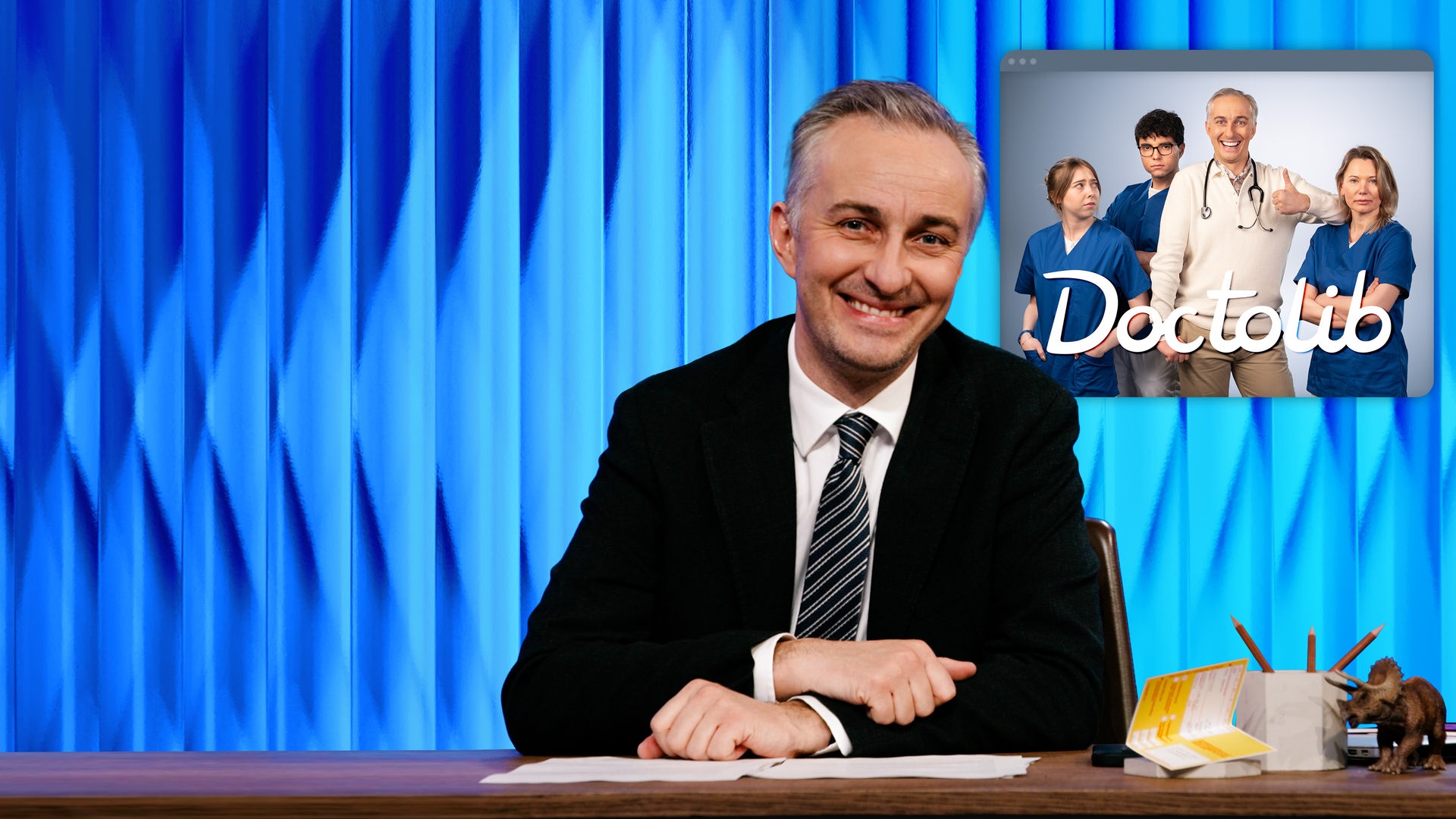 Jan Böhmermann sitzt am Schreibtisch. Rechts im Hintergrund ist eine Grafik zu sehen, in der er mit einem Stethoskop um den Hals und einer Daumen-hoch-Geste inmitten einer Gruppe medizinischer Fachkräfte steht. Am unteren Bildrand steht "Doctolib".