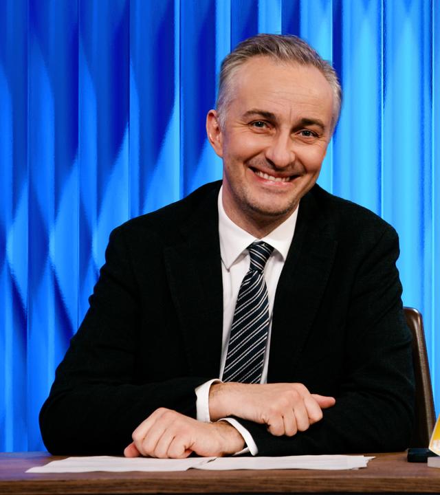 Jan Böhmermann sitzt am Schreibtisch. Rechts im Hintergrund ist eine Grafik zu sehen, in der er mit einem Stethoskop um den Hals und einer Daumen-hoch-Geste inmitten einer Gruppe medizinischer Fachkräfte steht. Am unteren Bildrand steht "Doctolib".