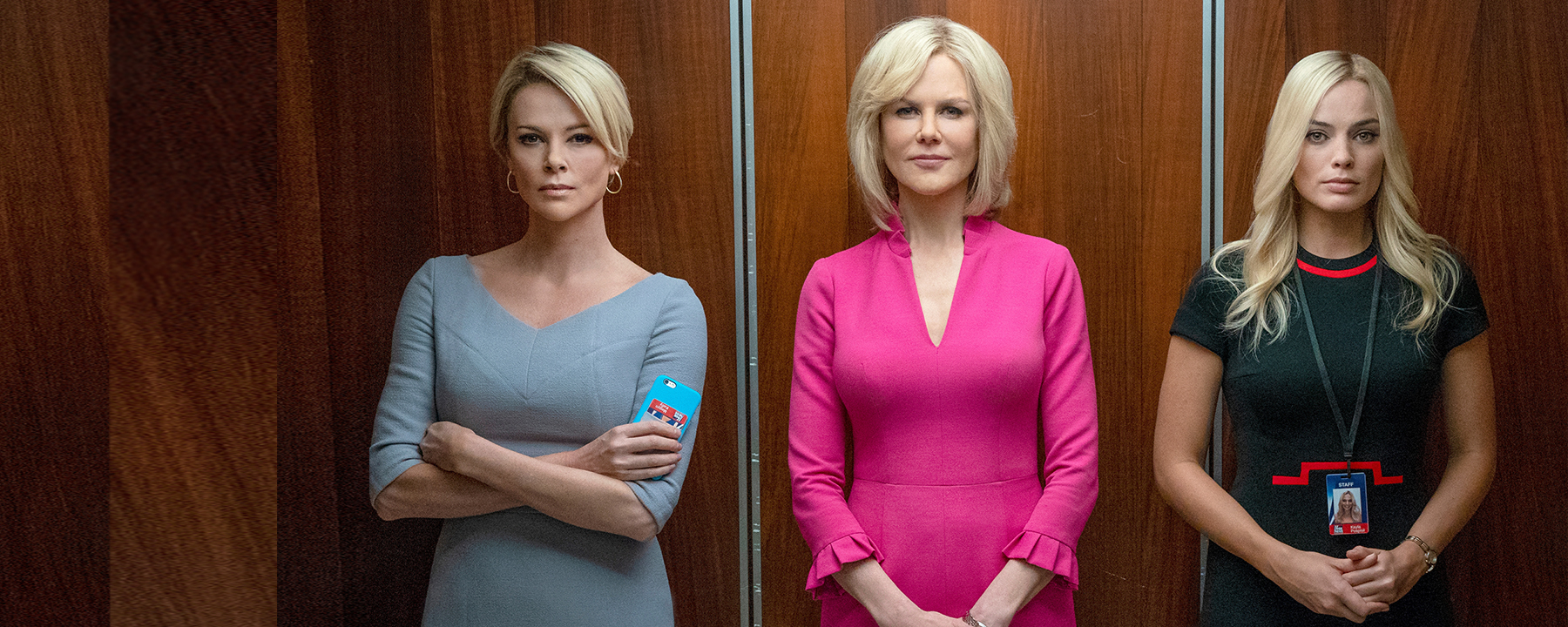 Drei Frauen in Etuikleidern stehen nebeneinander in einem offenen Fahrstuhl und blicken den Betrachter an. Die Frau in der Mitte, Gretchen Carlson (Nicole Kidman) trägt ein auffälliges, pinkfarbenes Kleid.