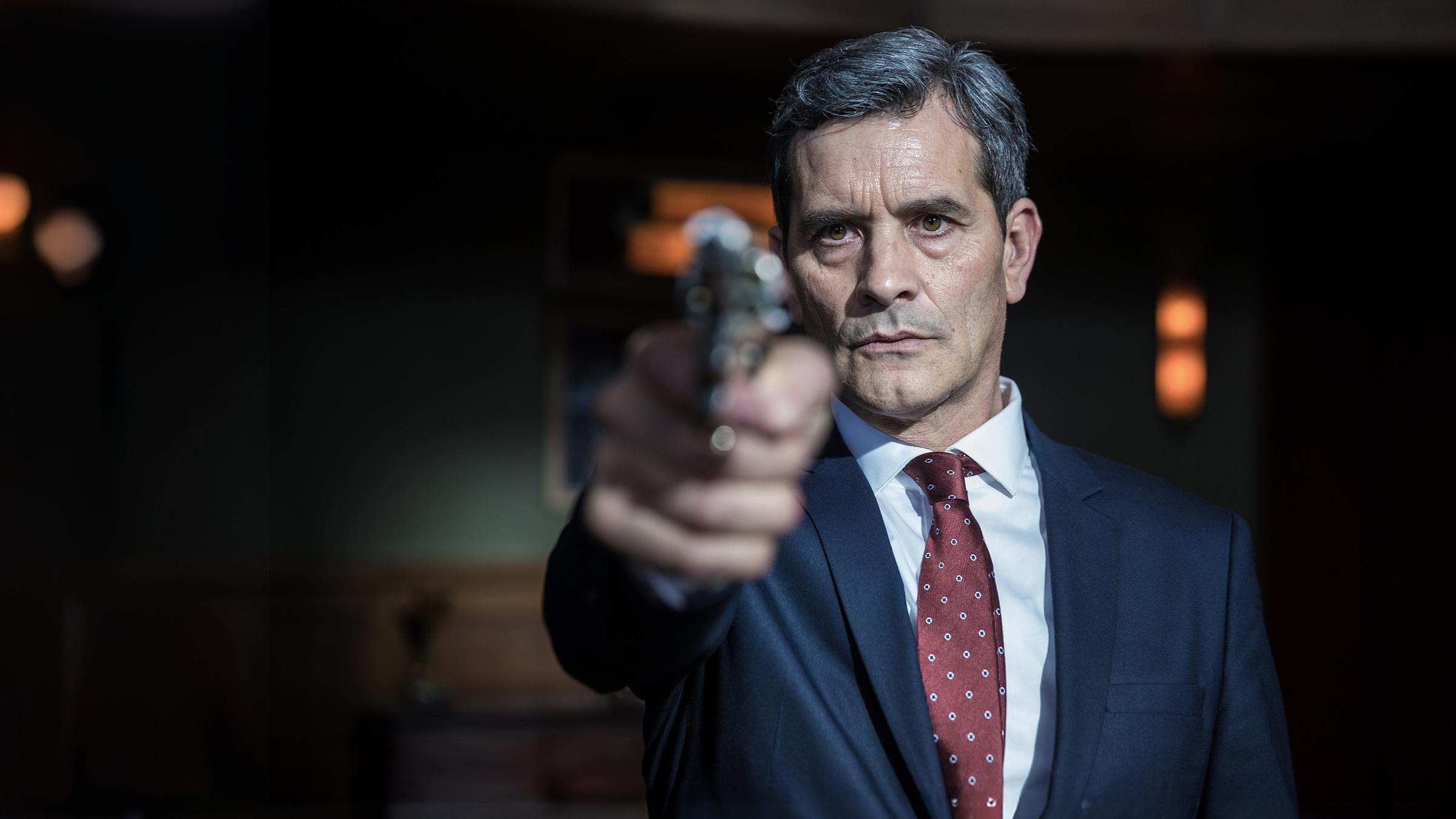 Der Premierminister (Koen De Bouw) in Nahaufnahme. Er hält eine Waffe in der Hand und richtet sie auf die Kamera.