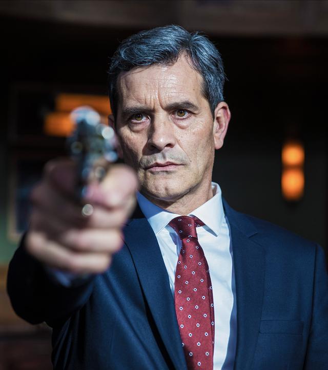 Ein Mann in einem blauen Anzug hat eine Waffe im Anschlag