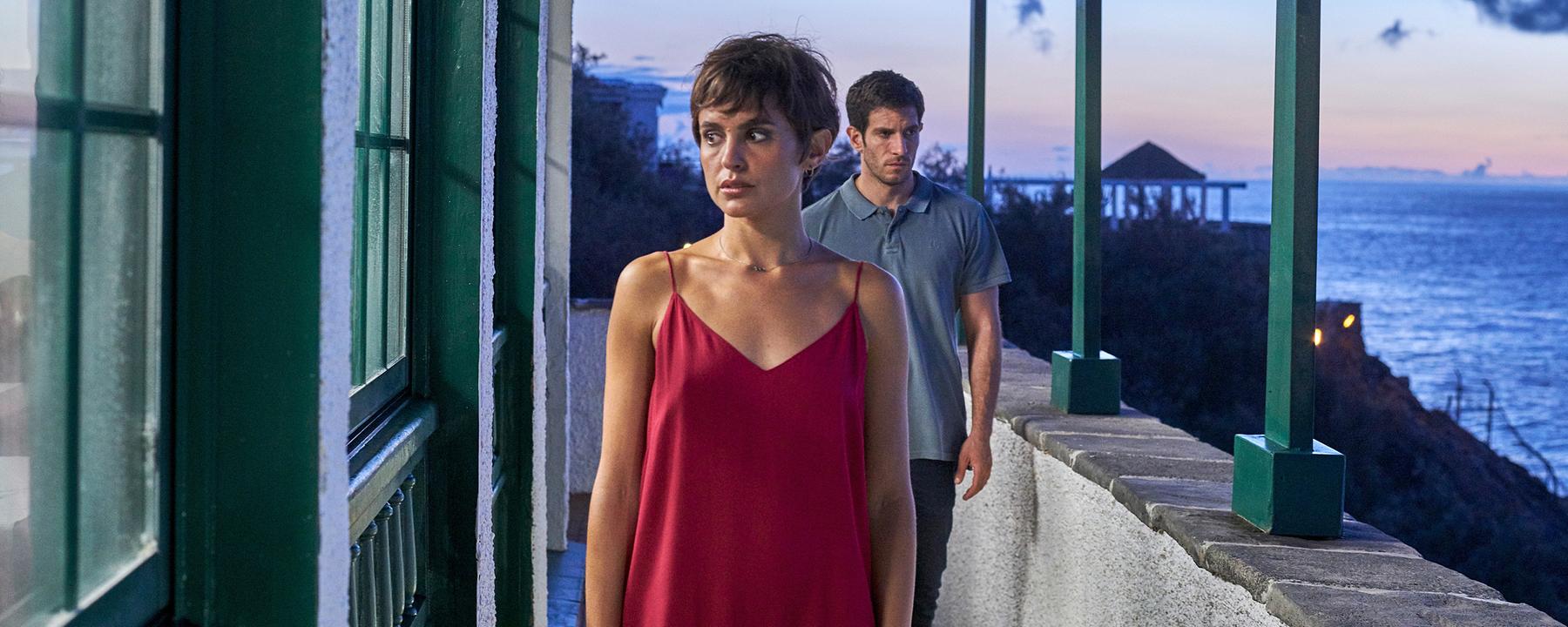 Ruth Anglada (Verónica Echegui) steht auf einem Balkon mit Meeresblick, hinter ihr nähert sich Sargento Bevilacqua (Quim Gutiérrez).