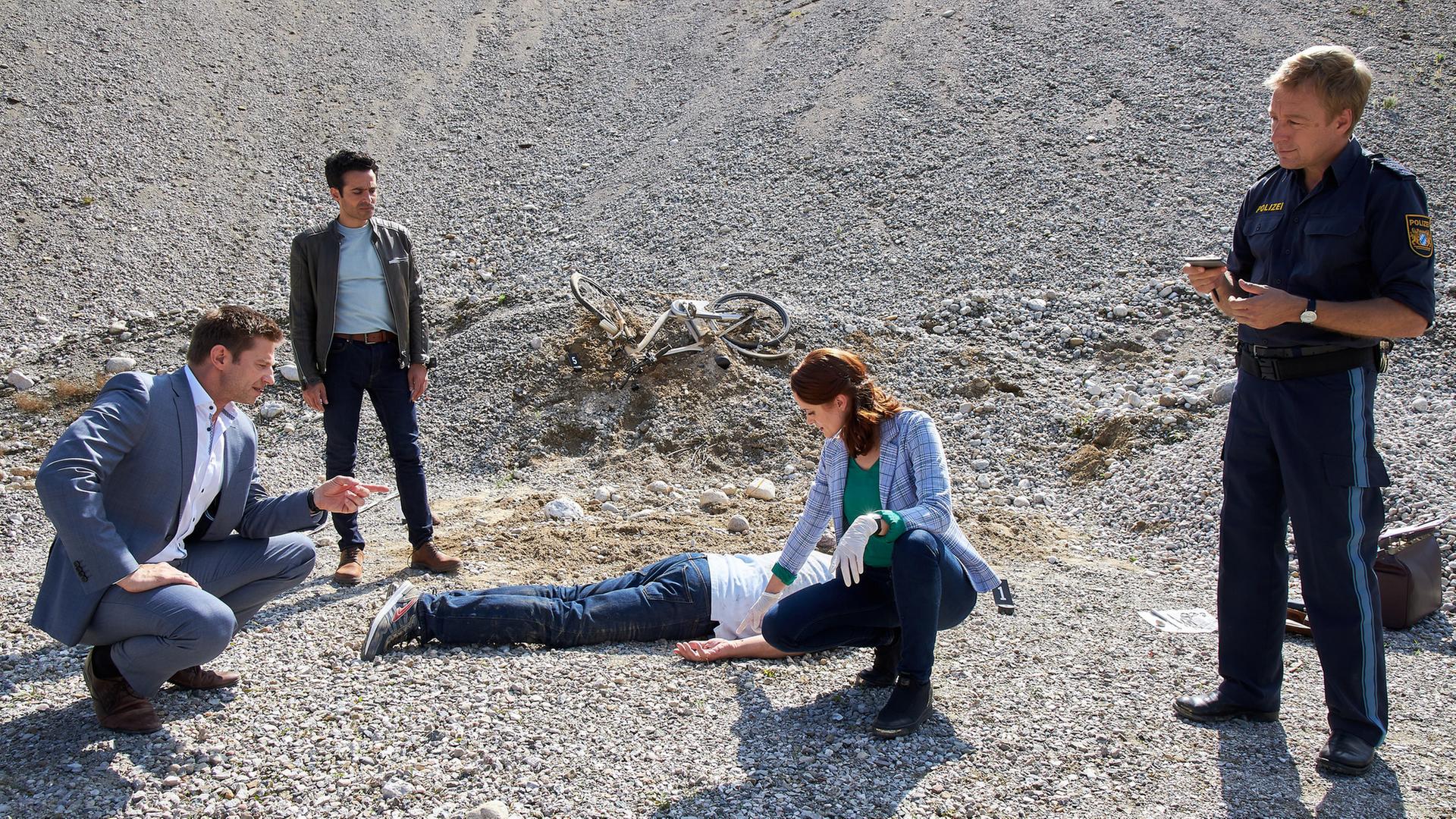 "Die Rosenheim-Cops - Mord aus der Luft": Kommissar Hansen (Igor Jeftić) hockt in einer Kiesgrube und spricht mit Pathologin Mai (Sina Wilke), die neben dem Opfer hockt und auf dessen Hand deutet. Neben ihnen stehen Kommissar Kaya (Baran Hêvî) und Polizeihauptmeister Mohr (Max Müller). Im Hintergrund liegt ein verbogenes Fahrrad am Hang.