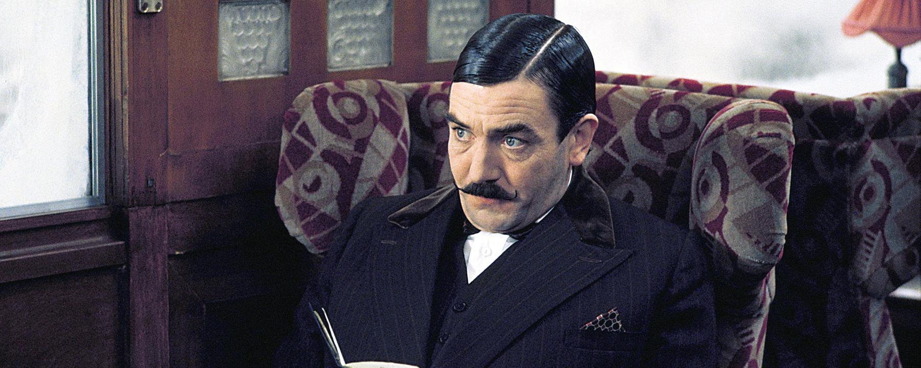 Hercule Poirot (Albert Finney), mit Schnurrbart und dunklem Anzug, sitzt in einem Zugabteil an einem Tisch.
