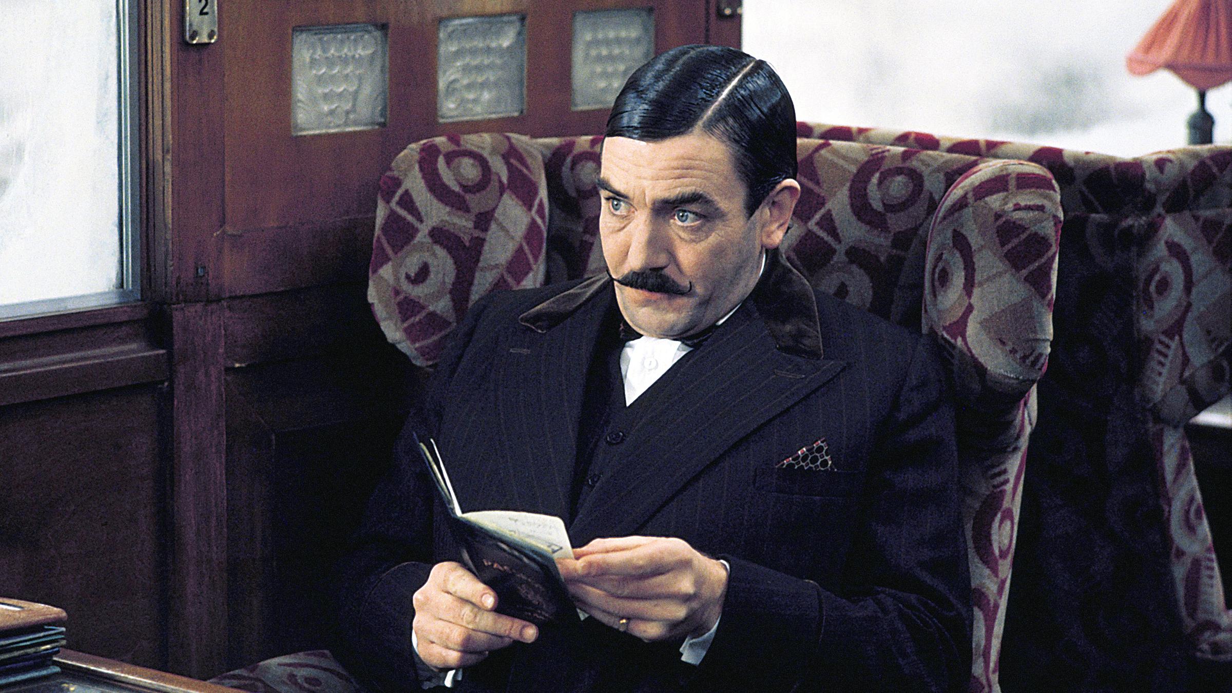 Hercule Poirot (Albert Finney), mit Schnurrbart und dunklem Anzug, sitzt in einem Zugabteil an einem Tisch.
