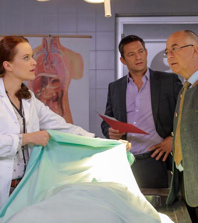 "Die Rosenheim-Cops - Mord mit Verspätung": Pathologin Sandra Mai (Sina Wilke) legt ein Tuch über eine Leiche, die vor ihr auf dem Tisch liegt. Kommissar Hansen (Igor Jeftic) und Polizeichef Achtziger (Alexander Duda) stehen daneben.