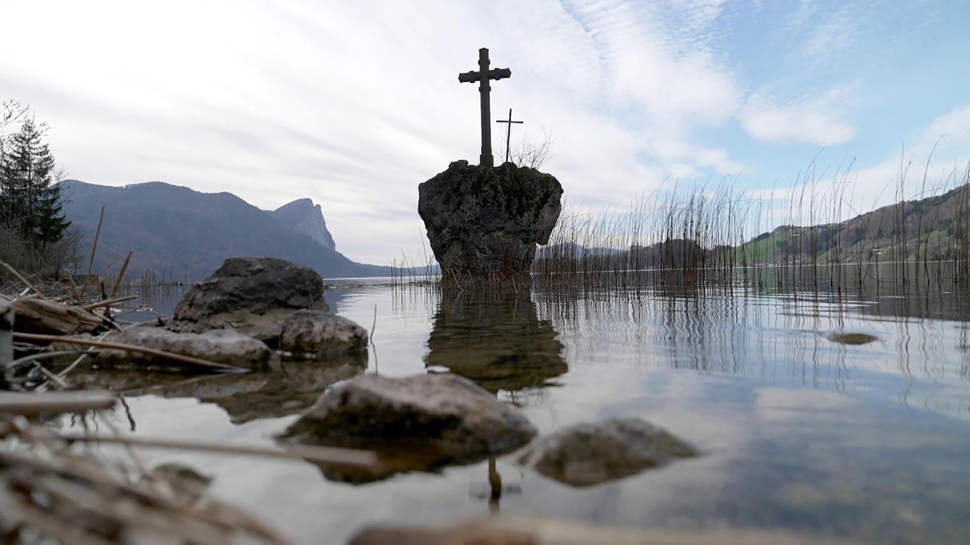 Das Bild zeigt eine ruhige Seenlandschaft mit einer Wasserfläche, die teilweise mit Schilfrohr und kleinen Steinen gesäumt ist. In der Mitte des Bildes steht ein kleiner, isolierter Felsen, auf dem zwei Holzkreuze platziert sind. Der Hintergrund besteht aus sanften Hügeln, die durch ihre bewaldeten Flächen und einige freie Wiesen gekennzeichnet sind. Der Himmel ist bewölkt, mit sanften, grauen und blauen Farbtönen. Die Szenerie vermittelt eine friedliche, aber auch melancholische Stimmung, die auf die Tiefe des dargestellten Themas hinweist.