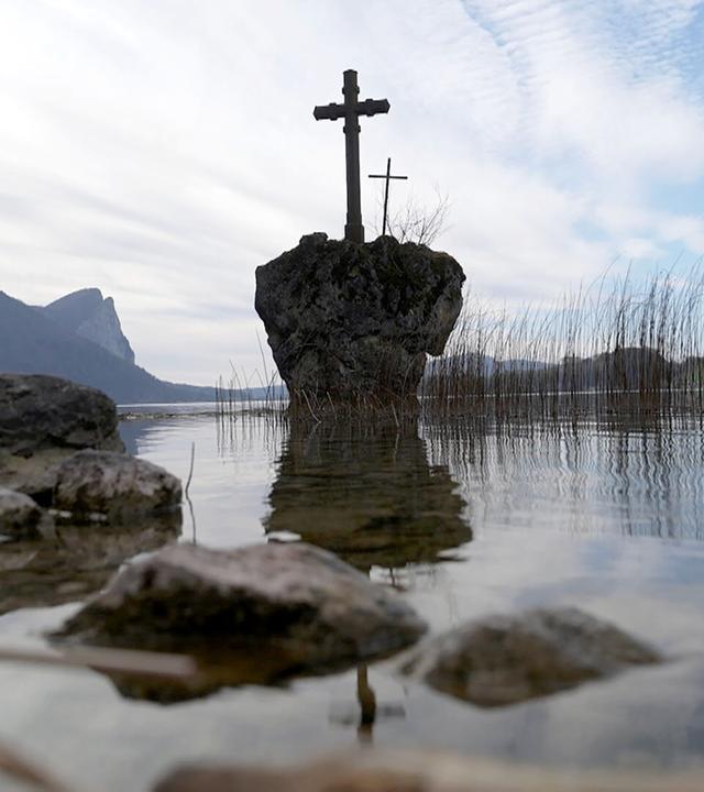 Das Bild zeigt eine ruhige Seenlandschaft mit einer Wasserfläche, die teilweise mit Schilfrohr und kleinen Steinen gesäumt ist. In der Mitte des Bildes steht ein kleiner, isolierter Felsen, auf dem zwei Holzkreuze platziert sind. Der Hintergrund besteht aus sanften Hügeln, die durch ihre bewaldeten Flächen und einige freie Wiesen gekennzeichnet sind. Der Himmel ist bewölkt, mit sanften, grauen und blauen Farbtönen. Die Szenerie vermittelt eine friedliche, aber auch melancholische Stimmung, die auf die Tiefe des dargestellten Themas hinweist.
