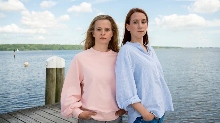 "Mordsschwestern - Verbrechen ist Familiensache: Schwarzer Fisch": Feli (Caroline Hanke) und Viktoria (Lena Dörrie) stehen auf einem Steg und schauen in Richtung Kamera. Im Hintergrund sieht man das Meer.