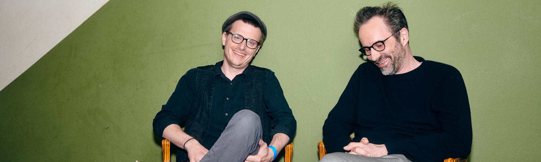 In einem Hausflur vor einer grünen Wand sitzen Moritz Neumeier und Kurt Krömer auf alten Lederstühlen. Neumeier lacht, Krömer schaut auf den Boden und lacht.