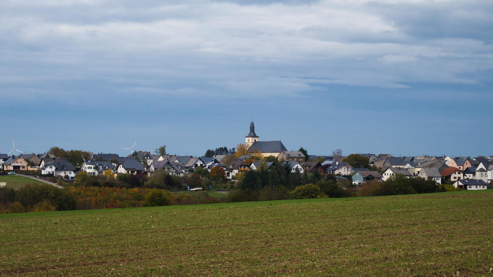 Blick auf ein kleines Dorf mit Kirche hinter einem Feld, auf dem es grün sprießt. Darüber blauer Himmel.