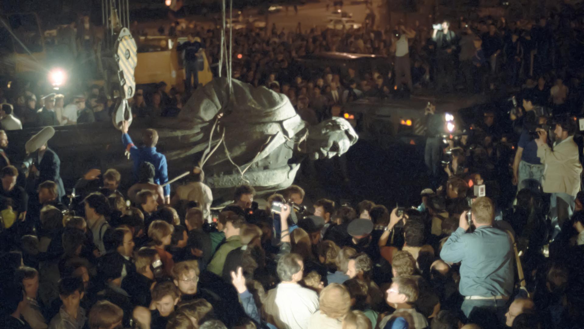 Szene bei Nacht: Eine Menschenmenge schaut zu und fotografiert, wie eine riesige sowjetische Statue von einem Mann mit Seilen vom Sockel heruntergeholt und abtransportiert wird.