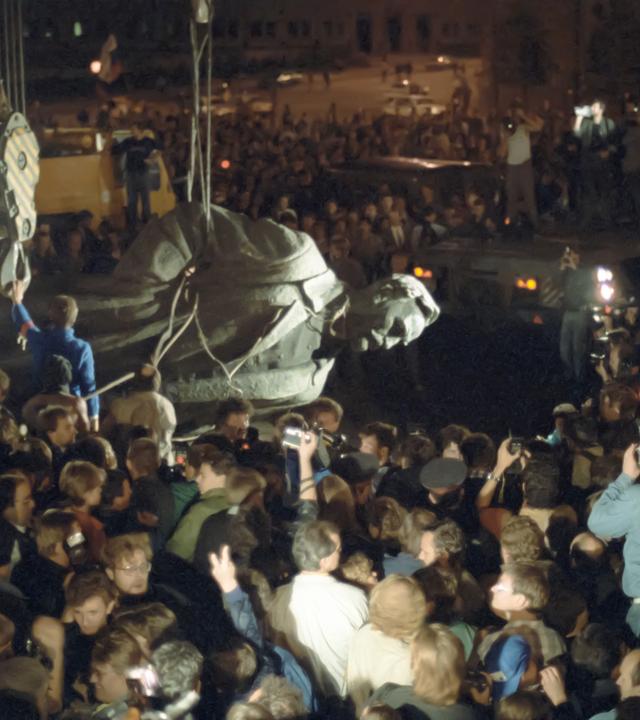 Szene bei Nacht: Eine Menschenmenge schaut zu und fotografiert, wie eine riesige sowjetische Statue von einem Mann mit Seilen vom Sockel heruntergeholt und abtransportiert wird.