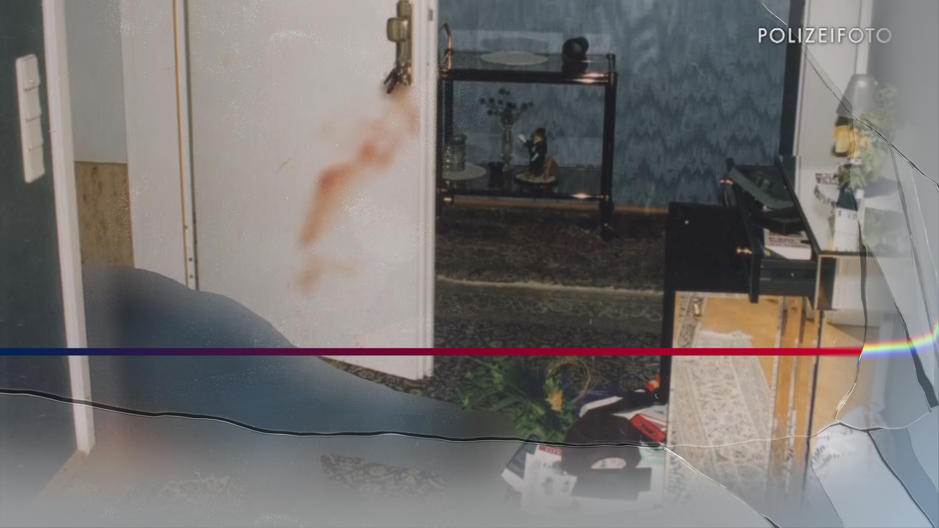 Bild aus der Polizeiakte. Beine eine Leiche (unkenntlich) liegen auf einer Türschwelle. An der Tür sind Bluspuren zu erkennen. Ein Schlüssel steckt im Schloss.