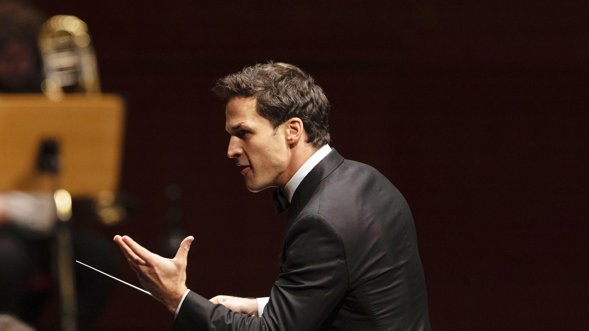 Das Bild zeigt einen männlichen Dirigenten, der sich zur Seite wendet. Er trägt einen schwarzen Frack und hat ein helles Hemd mit Kläppchenkragen an. Sein Gesichtsausdruck ist intensiv und engagiert, während er mit einer Hand gestikuliert. In der anderen Hand hält er einen Dirigentenstab, mit dem er die musikalischen Anweisungen gibt. Im Hintergrund sind unscharfe Konturen von Musikinstrumenten und Personen zu erkennen, die Teil des Orchesters sind. Die Szene vermittelt eine festliche und konzentrierte Atmosphäre, die für ein klassisches Konzert typisch ist.
