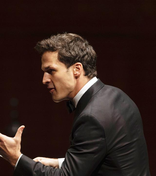 Das Bild zeigt einen männlichen Dirigenten, der sich zur Seite wendet. Er trägt einen schwarzen Frack und hat ein helles Hemd mit Kläppchenkragen an. Sein Gesichtsausdruck ist intensiv und engagiert, während er mit einer Hand gestikuliert. In der anderen Hand hält er einen Dirigentenstab, mit dem er die musikalischen Anweisungen gibt. Im Hintergrund sind unscharfe Konturen von Musikinstrumenten und Personen zu erkennen, die Teil des Orchesters sind. Die Szene vermittelt eine festliche und konzentrierte Atmosphäre, die für ein klassisches Konzert typisch ist.