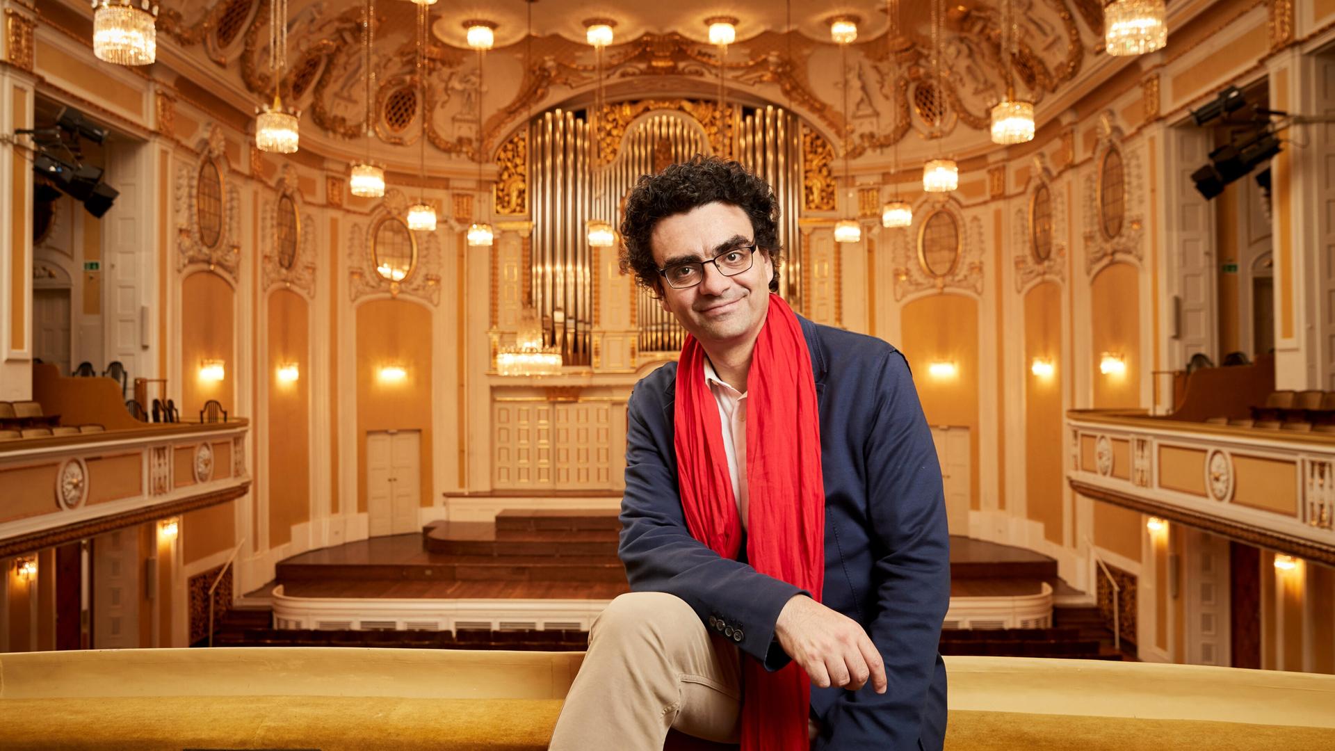 Das Bild zeigt einen Mann, der entspannt auf einer Geländerbrüstung in einem prunkvollen Konzertsaal sitzt. Er trägt ein blaues Sakko, ein helles Hemd und einen auffälligen roten Schal. Der Mann hat lockiges, dunkles Haar und eine Brille.   Im Hintergrund sind die charakteristischen Merkmale des Konzertsaals zu sehen: hohe, mit kunstvollen Verzierungen dekorierte Wände in sanften Farbtönen, mehrere raffinierte Kronleuchter, die den Raum beleuchten, und ein großes, aufwendiges Orgelinstrument an der Wand. Die Bühne ist mit hellem Holz ausgelegt und hat Treppenstufen, die ins Publikum führen. Die gesamte Atmosphäre vermittelt einen eleganten und festlichen Eindruck, passend zu einem Musikfestival.