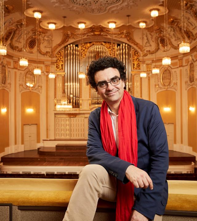 Das Bild zeigt einen Mann, der entspannt auf einer Geländerbrüstung in einem prunkvollen Konzertsaal sitzt. Er trägt ein blaues Sakko, ein helles Hemd und einen auffälligen roten Schal. Der Mann hat lockiges, dunkles Haar und eine Brille.   Im Hintergrund sind die charakteristischen Merkmale des Konzertsaals zu sehen: hohe, mit kunstvollen Verzierungen dekorierte Wände in sanften Farbtönen, mehrere raffinierte Kronleuchter, die den Raum beleuchten, und ein großes, aufwendiges Orgelinstrument an der Wand. Die Bühne ist mit hellem Holz ausgelegt und hat Treppenstufen, die ins Publikum führen. Die gesamte Atmosphäre vermittelt einen eleganten und festlichen Eindruck, passend zu einem Musikfestival.