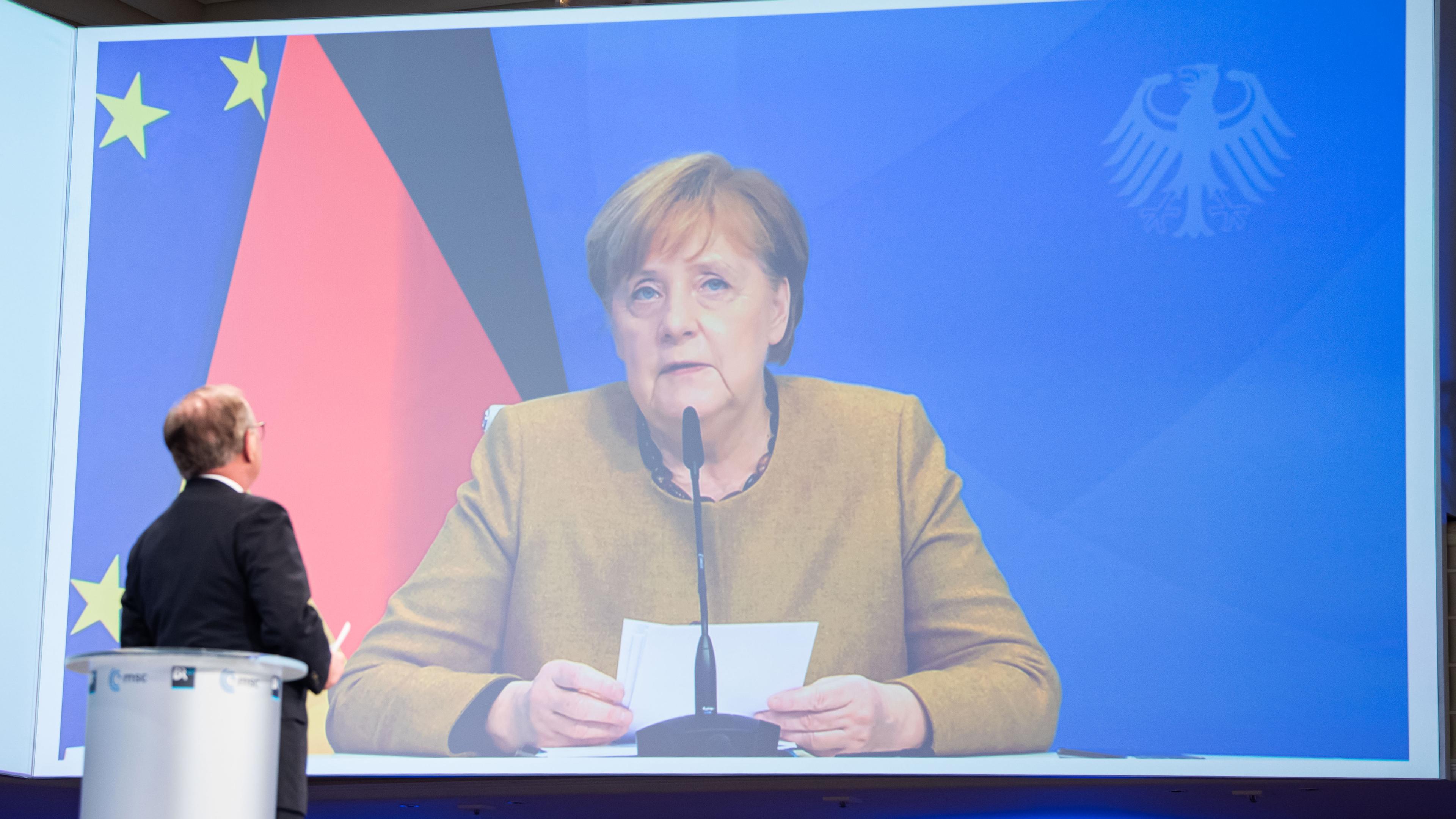 Zu sehen ist Angela Merkel auf einer großen Leinwand, wie sie sitzend eine Rede hält. Hinter ihr sieht man die deutsche Flagge.