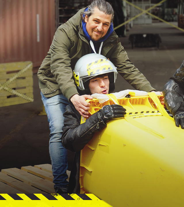 Tommy hält eine waagerechte Mülltonne, in der Felix mit einem Helm sitzt. Kenneth trägt ebenfalls einen Helm und schiebt die Mülltonne von vorne an.