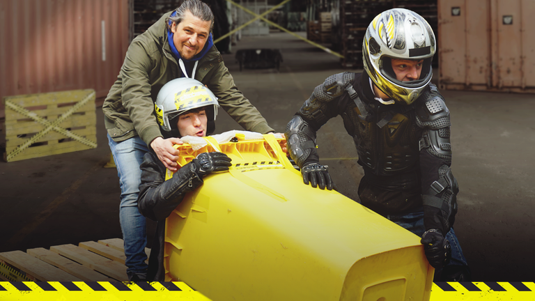 Tommy hält eine waagerechte Mülltonne, in der Felix mit einem Helm sitzt. Kenneth trägt ebenfalls einen Helm und schiebt die Mülltonne von vorne an.