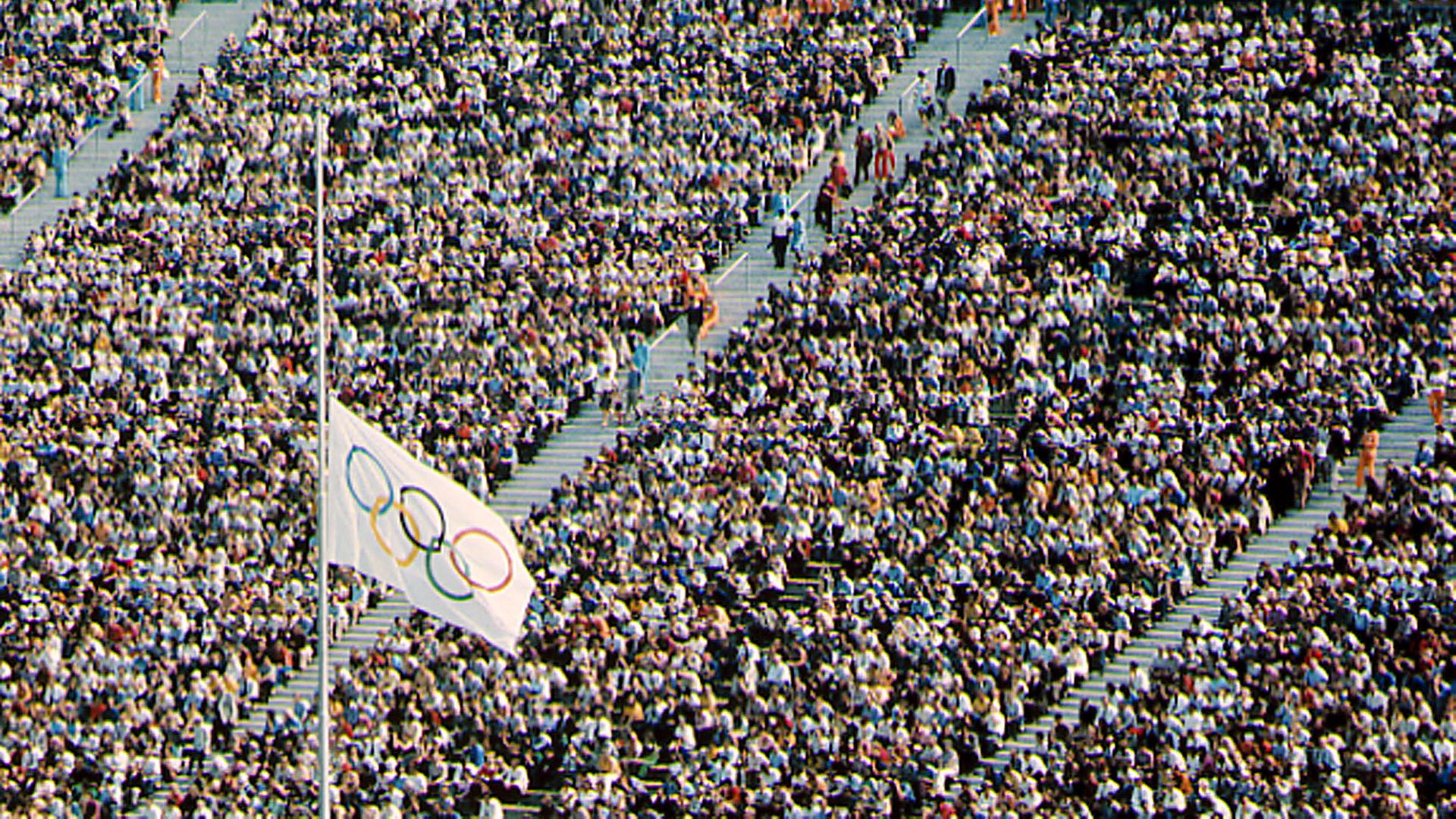 Menschen nehmen im Münchner Olympiastadion an der Gedenkfeier teil. Die Olympiafahne steht auf Halbmast.
