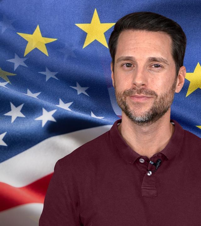 Mirko Drotschmann ist zu sehen, vor einem Hintergrund auf dem sowohl die US-Flagge als auch die EU-Flagge zu erkennen sind