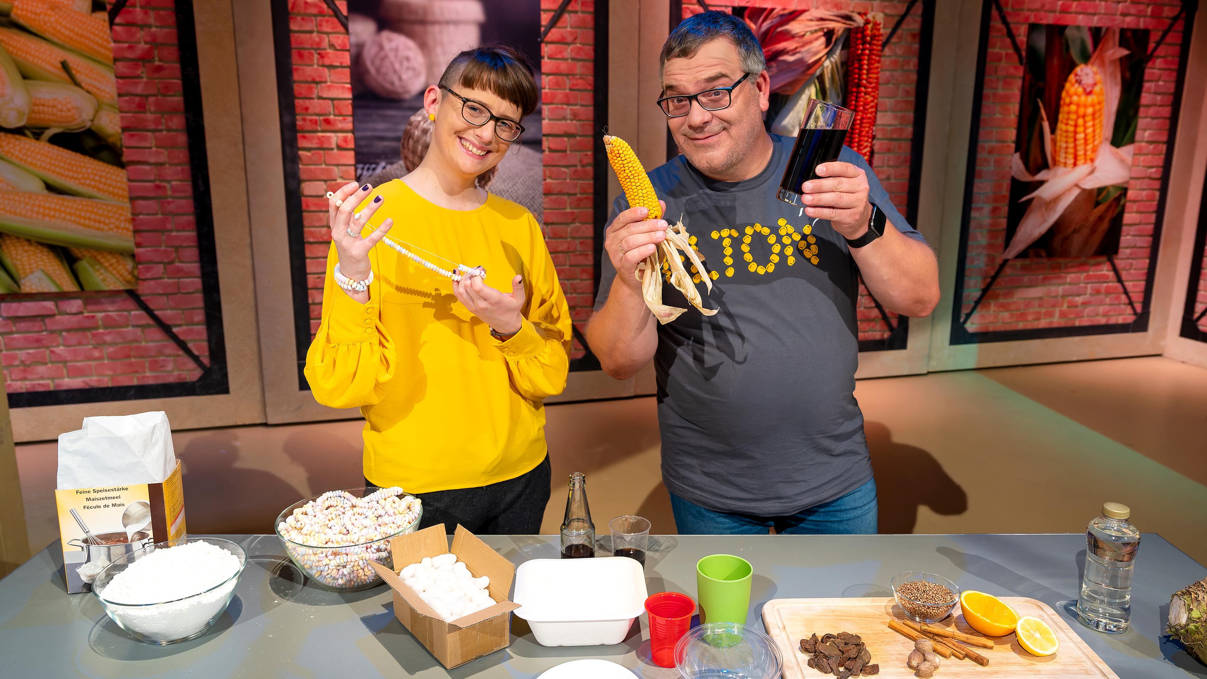 1, 2 oder 3: Moderator Elton steht mit Chemikerin und Princess of Science Patrizia Thoma im "1, 2 oder 3"-Studio. Vor ihnen steht ein Tisch mit verschiedenen maishaltigen Gegenständen und Lebensmitteln.