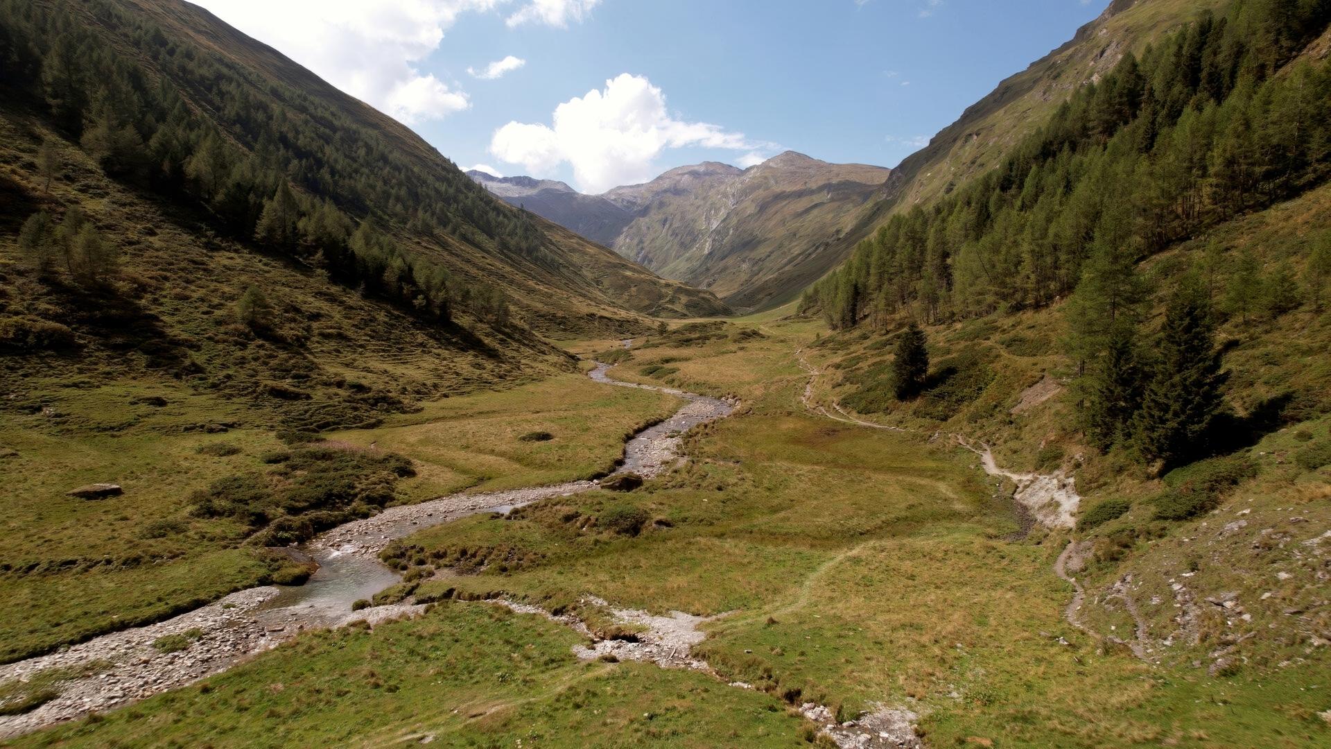 Das Bild zeigt eine weite, grüne Landschaft im Nationalpark Hohe Tauern in Österreich. In der Mitte schlängelt sich ein kleiner, klarer Bach durch die Wiesen. Umgeben ist die Szene von sanften Hügeln und steilen Bergen, die sich im Hintergrund erheben und zum Teil mit Gras und kleinen Bäumen bewachsen sind. Der Himmel ist teilweise bewölkt, es sind jedoch auch sonnige Abschnitte sichtbar. Die gesamte Umgebung strahlt Ruhe und Naturverbundenheit aus.