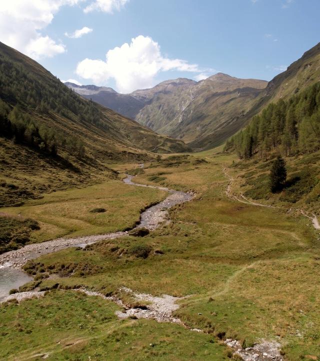 Das Bild zeigt eine weite, grüne Landschaft im Nationalpark Hohe Tauern in Österreich. In der Mitte schlängelt sich ein kleiner, klarer Bach durch die Wiesen. Umgeben ist die Szene von sanften Hügeln und steilen Bergen, die sich im Hintergrund erheben und zum Teil mit Gras und kleinen Bäumen bewachsen sind. Der Himmel ist teilweise bewölkt, es sind jedoch auch sonnige Abschnitte sichtbar. Die gesamte Umgebung strahlt Ruhe und Naturverbundenheit aus.