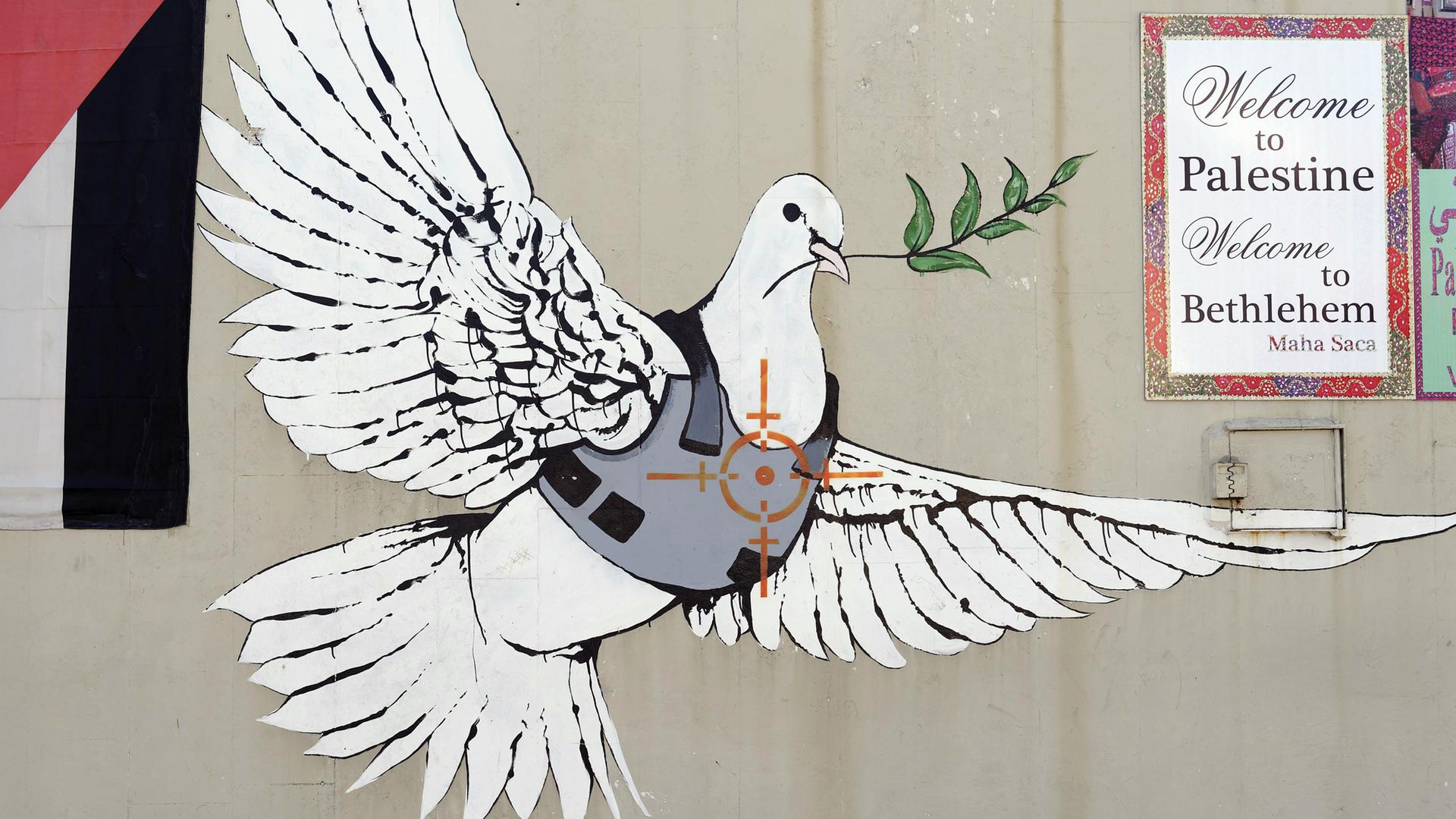 Auf eine Wand ist eine Friedenstaube mit Schutzweste und Zielkreuz auf der Brust gemalt, die einen Olivenzweig im Schnabel trägt.