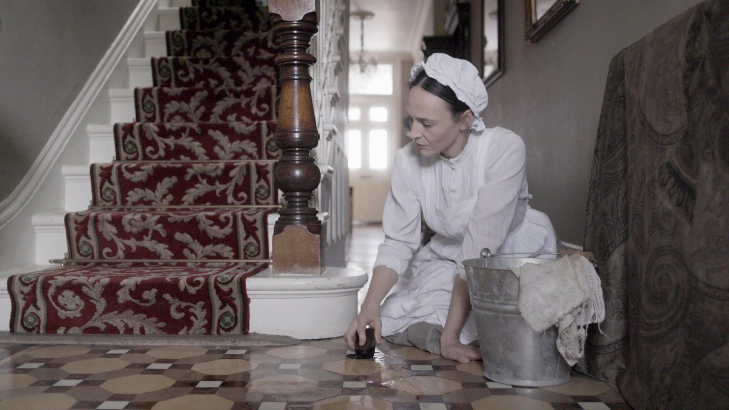"Murder Maps: Das irische Dienstmädchen": Spielszene: Kate Webster (Darstellerin unbekannt) kniet beim Putzen auf dem Boden neben einer Treppe eines herrschaftlichen Hauses. Neben ihr steht ein Putzeimer.