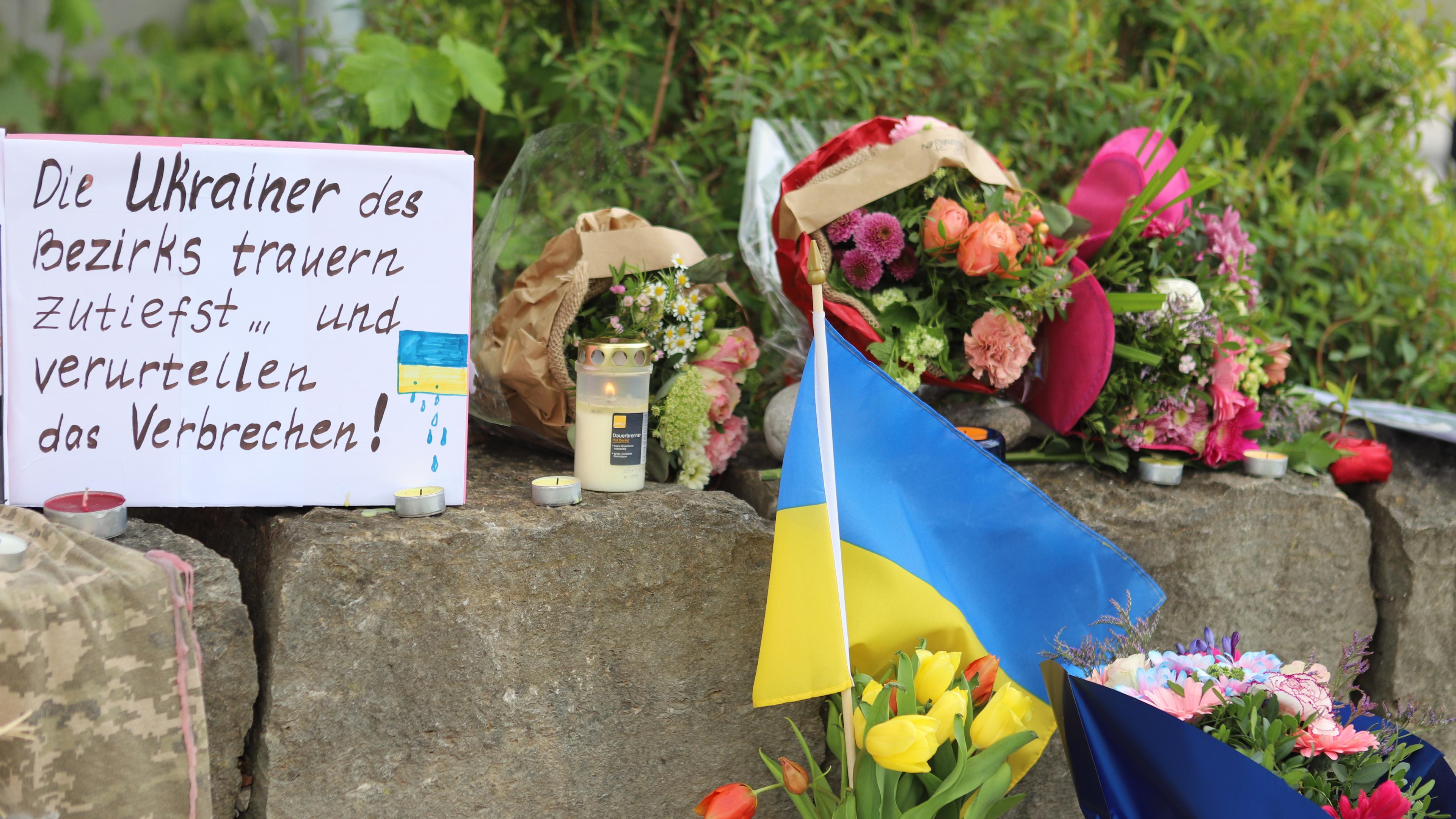 Blumen, Kerzen, ein Schild mit Worten der Trauer und die ukrainische Flagge liegen auf einer Steinmauer
