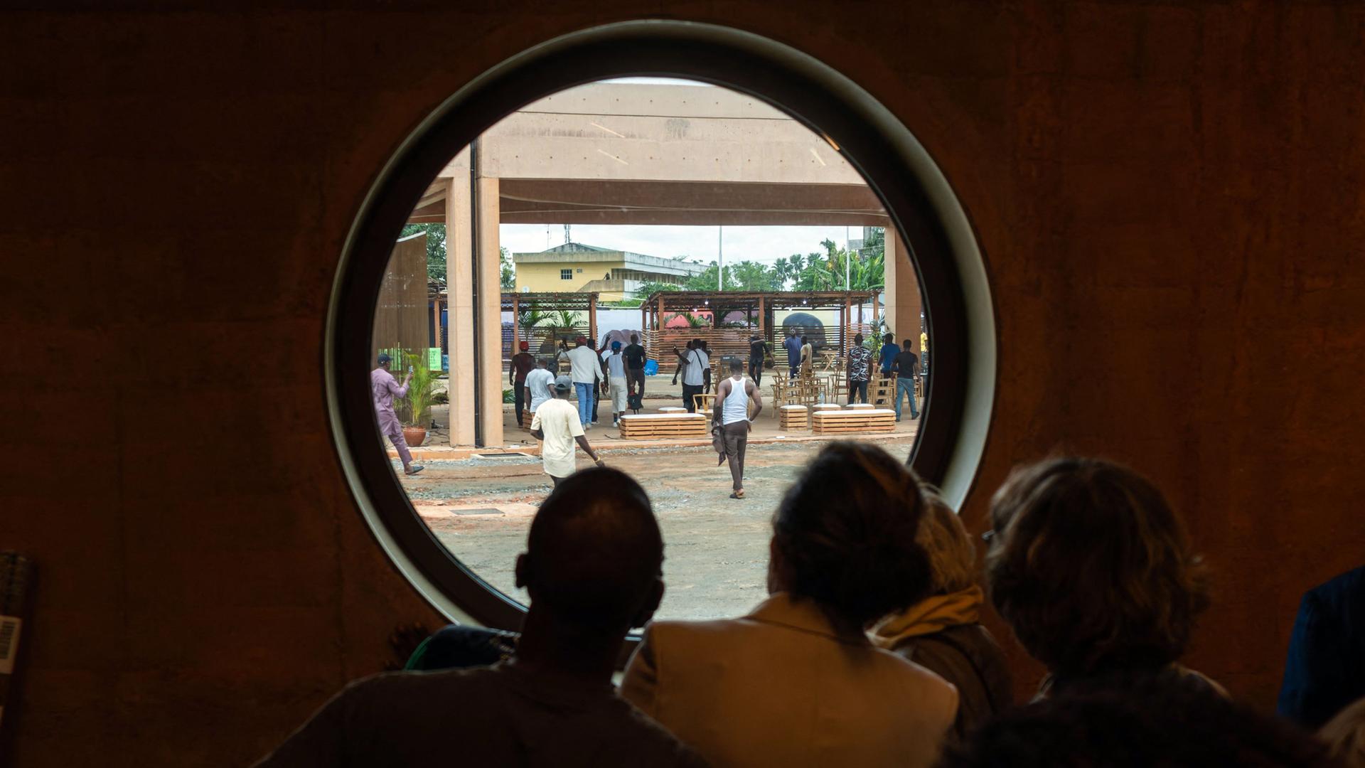 Rückansicht mehrerer Menschen, die durch ein großes Bullauge nach draußen schauen, wo protestierende Männer herumlaufen.
