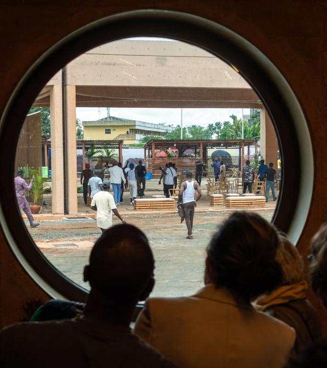 Rückansicht mehrerer Menschen, die durch ein großes Bullauge nach draußen schauen, wo protestierende Männer herumlaufen.