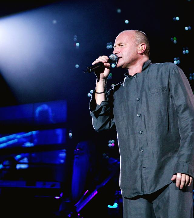 Phil Collins singt auf der Bühne in ein Mikro.
